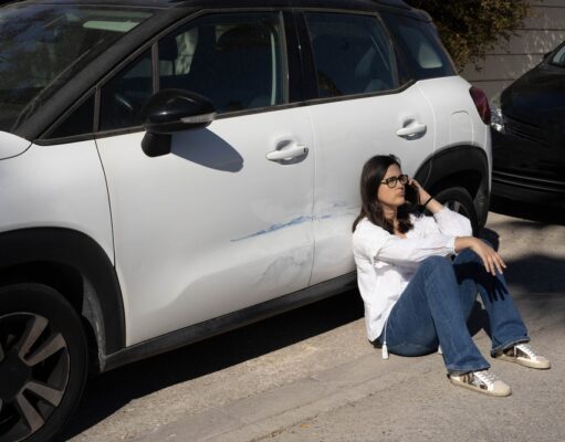 Vandalizmus na aute – ako postupovať po poškodení vozidla? Žena sedí pri poškriabanom aute a telefonuje, rieši vandalizmus na vozidle.