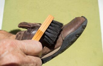 Ako sa starať o turistické topánky, aby vydržali roky? Detail ruky čistiacej koženú turistickú topánku kefou na zelenom pozadí.